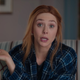 Wanda Maximoff, in a blue plaid shirt, sits on a striped couch and talks directly into the camera.